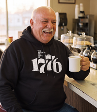 Man wearing The Answer To 1984 Is 1776 Hoodie in black at a diner counter