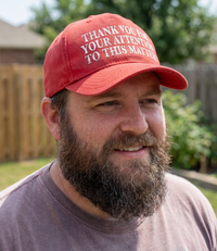 The Alex Jones Store - Man wearing red 'Thank You For Your Attention To This Matter' trucker hat with white embroidered text, outdoors in a backyard