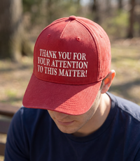 The Alex Jones Store - Close-up of red curved-brim cap with white 'Thank You For Your Attention To This Matter!' embroidery worn outdoors