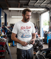 The Alex Jones Store - Bearded mechanic in auto shop wearing white Fed Slop t-shirt featuring cartoon sheep eating from mainstream media bowl