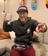 Alex Jones Naturals - Man in black Anchor cap and hoodie holding jar of Optimal Human Superfood Greens while seated on a couch pointing at camera