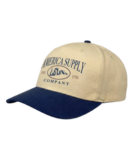 The Alex Jones Store - Tan and navy America Supply Co. 'Est. 1776' Trucker Hat with rattlesnake oval logo, side-angle showing curved navy bill