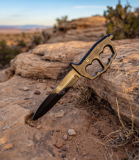 The Alex Jones Store - Brass-finished Ave Christus Rex Trench War Dagger with knuckle-guard handle staged on desert sandstone at sunset