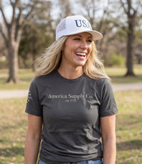 The Alex Jones Store - Blonde woman laughing outdoors in gray America Supply Co. 'Est. 1776' tee and white USA trucker hat