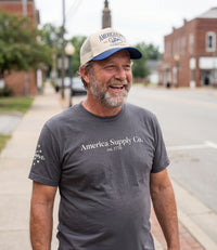 The Alex Jones Store - Bearded man smiling on sidewalk wearing gray America Supply Co. 'Est. 1776' tee and America Supply Co. trucker hat