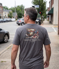The Alex Jones Store - Man walking down street wearing gray America Supply Co. 'Est. 1776' tee, back view showing drum and flag print