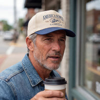 The Alex Jones Store - Man in cream and navy America Supply Co trucker hat with 'Est. 1776' rattlesnake logo, holding coffee on a city street