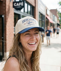The Alex Jones Store - Smiling woman wearing tan and navy America Supply Co. 'Est. 1776' Trucker Hat with rattlesnake logo on city sidewalk