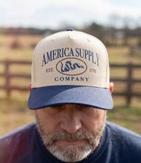The Alex Jones Store - Bearded man wearing tan and navy America Supply Co. 'Est. 1776' Trucker Hat with rattlesnake logo, rural fence backdrop