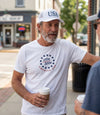 The Alex Jones Store - Man wearing white America 250 t-shirt with navy '1776/2026' star wreath emblem on chest and white USA cap, outdoor lifestyle shot
