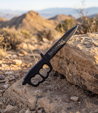 The Alex Jones Store - Black Ave Christus Rex Trench War Dagger with 'Ave Christus Rex' Gothic etched blade resting on a desert mountain rock