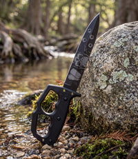 The Alex Jones Store - Liberty or Death Trench War Dagger leaning against a granite boulder beside a Hill Country creek