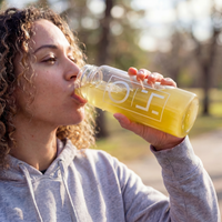 Alex Jones Naturals - Optimal Human Hydration Drink Mix with French Sea Salt — Woman drinking yellow Optimal Human hydration drink from a glass OH branded bottle outdoors in soft natural sunlight
