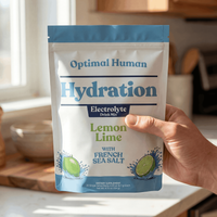 Alex Jones Naturals - Hand holding Optimal Human Hydration Lemon Lime electrolyte drink mix pouch with French sea salt in a sunlit kitchen